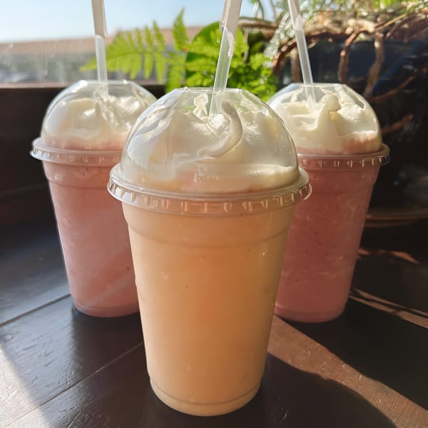 Three vibrant smoothies with whipped cream and straws, two pink and one orange, on a dark wooden surface.
