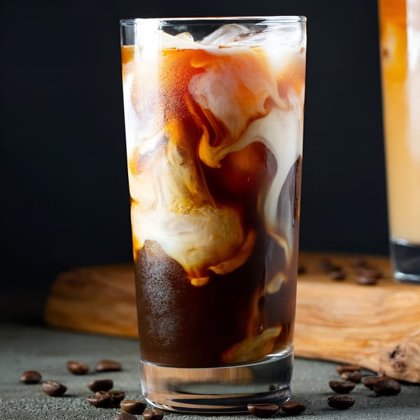 Iced coffee with cream swirling into dark coffee, surrounded by coffee beans on a dark surface.